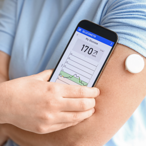 picture of guy holding phone with an app for blood sugar monitoring next to his arm with a glucometer - for the blog Benefits of Weightlifting on Blood Sugar