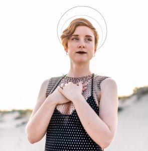 A picture of Lorna Stell, an intuitive coach and healing guide, on a beach with a halo headband and her hands clasped at her heart, for the podcast interview with Hope Health and Wellness Advocacy